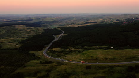 Aerial-drone-landscape-view-over-a-road-winding-through-a-Romanian-forest-and-countryside,-at-sunset