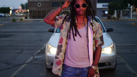 African-American-man-with-dreadlocks-wearing-dark-circle-sunglasses-in-front-of-car-headlights-and-adjusting-his-hair-as-camera-moves-in-towards-him