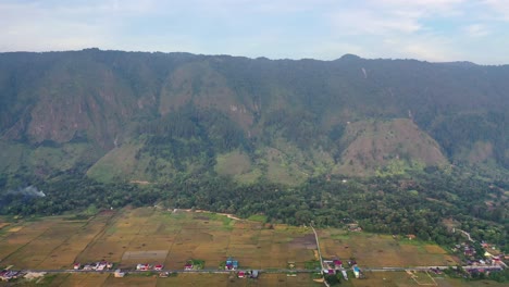 Backwards-aerial-shot-of-village-and-fields-below-hills-on-Samosir-Island-in-Lake-Toba-in-North-Sumatra,-Indonesia