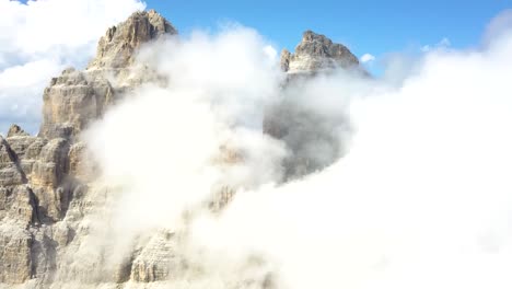 Ascending-Aerial-of-a-Mountain-Peak-on-a-sunny-day,-with-fluffy-white-clouds-covering-the-top-at-National-Park-Tre-Cime-di-Lavaredo,-Alps,-Dolomites,-Italy