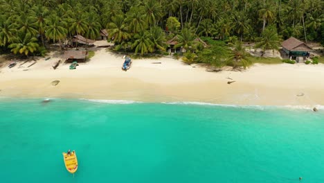 Drone-flying-away-from-beach-with-boat-and-simple-huts-on-tropical-island-surrounded-by-amazing-turquoise-water-in-North-Sumatra,-Indonesia