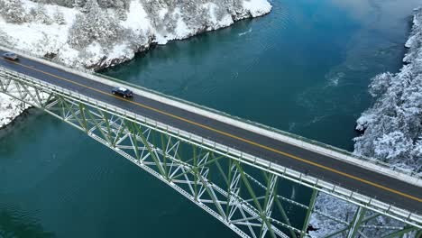 Close-up-aerial-view-of-a-car-driving-on-a-bridge-high-up-above-the-water-with-snow-everywhere