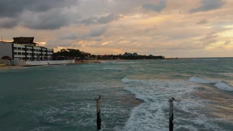 Flying-low-over-the-turquoise-blue-waters-of-the-Caribbean-sea-to-reveal-the-hotels-and-beachfront-of-playa-Paraiso-beach-in-Tulum-Mexico-on-a-cloudy-day-with-a-golden-hour-sunset-sky