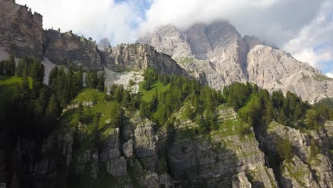 Ascending-aerial-up-a-green-cliff-side-on-a-mountain-peak-range-on-a-beautiful-sunny-day,-San-Vito-di-Cadore,-Tre-Cime,-Belluno,-Italy