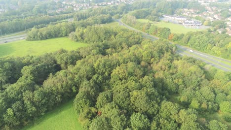 Green-UK-park-woodland-looking-down-aerial-view-above-motorway---suburban-neighbourhood
