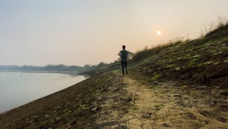 Rear-wide-view-of-man-jogging-on-rural-path-by-river-bed-in-Sylhet,-hazy-morning