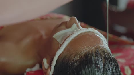 Ein-Stetiger-Ölstrahl-Fließt-über-Die-Stirn-Und-In-Die-Haare-Einer-Frau,-Die-Während-Eines-Ayurvedischen-Retreats-Eine-Entspannende-Shirodhara-Behandlung-Erhält