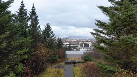 Drone-Flying-through-pine-tree-forest-pathway-to-Reykjavik-hallgrimskirkja,-on-hill,-then-Ascending-Above-Trees-to-Reveal-Hlíðarendi-and-City-of-Reykjavik,-Drone-Aerial