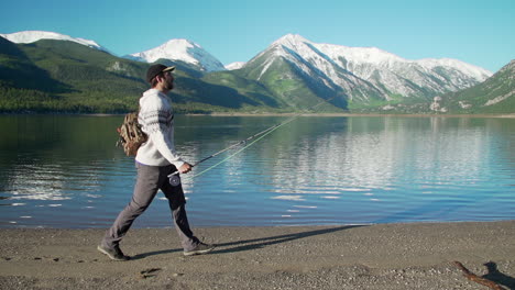 Wide-tracking-shot-of-a-fly-fisherman-walking-in-front-of-gorgeous,-snow-capped-mountains,-and-a-deep,-reflective-mountain-lake
