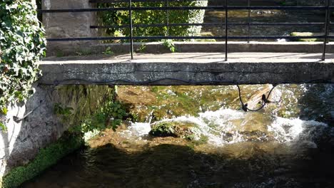 Fresh-stream-water-flowing-under-sunlit-stone-bridge-crossing-in-rural-countryside-village