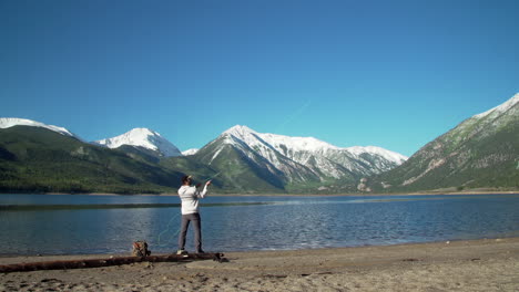 Wide-shot-of-a-fly-fisherman-casting-his-lure-into-a-deep-blue-lake-in-front-of-gorgeous,-snow-capped-mountains