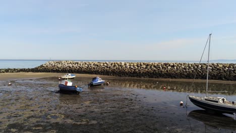 Aerial-low-angle-view-boats-in-shimmering-low-tide-sunny-warm-Rhos-on-Sea-seaside-sand-beach-marina-pill-back-reverse-right