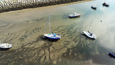 Aerial-birdseye-above-boats-in-shimmering-sunny-patterned-sand-beach-at-low-tide-marina-slow-dolly-left-pull-back