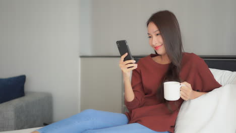 Asian-woman-with-a-beautiful-smile-is-using-her-mobile-phone-holding-a-cellphone-in-one-hand-and-typing-on-the-screen,-holding-a-cup-on-morning-coffee