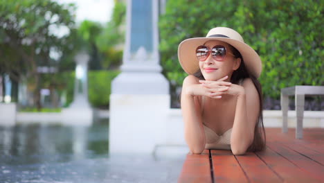 While-lying-prone-on-a-wooden-deck-surrounding-a-large-resort-pool,-a-young-woman-in-a-sun-hat-and-sunglasses-leaning-on-her-hands-smiles-as-she-looks-out-across-the-hotel-property