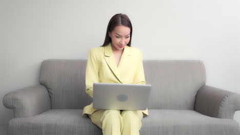 A-young-businesswoman-in-a-pale-yellow-business-suit-works-on-her-laptop-while-sitting-on-a-couch