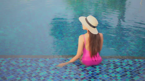 Woman-wearing-pink-bathing-suit-and-large-straw-hat-sitting-in-clear-warm-swimming-pool-with-decorative-tile
