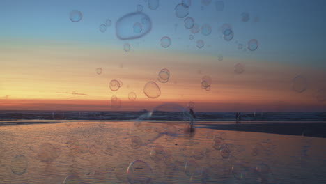 Glänzende-Seifenblasen,-Die-über-Den-Strand-In-Richtung-Feurigen-Sonnenuntergangshimmel-Mit-Orange-blauen-Farben-Schweben,-Zeitlupenweite-Aufnahme
