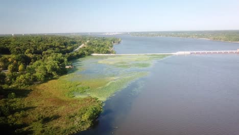 Aerial-view-of-upstream-backwater-of-Lock-and-Dam-14-on-Mississippi-River-with-dike,-waterlilies,-duckweed-and-trees-along-the-shore