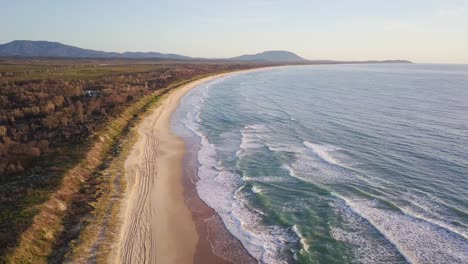 Aerial-view-of-a-secluded-beach-in-New-South-Wales,-Australia-with-long-and-calm-waves-crashing-the-coast-at-sunset