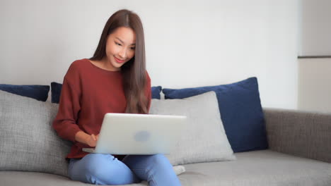Asian-woman-using-her-laptop-computer-sitting-on-the-bed,-she-is-typing-on-the-laptop-keyboard-and-suddenly-wins-the-prize,-lottery,-exited-face-expression,-raise-hands-up
