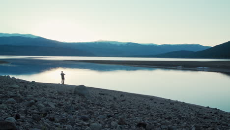 Wide-silhouette-shot-of-a-fisherman-casting-his-lure-into-a-still,-glassy,-lake-in-front-of-gorgeous,-snow-capped-mountains