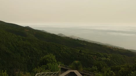 Luftaufnahme-Von-Einem-Bewaldeten-Berghang-Nach-Vorne,-Im-Hintergrund-Eine-Stadt-Und-Ein-Wolkenmeer
