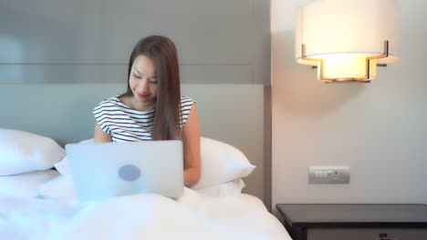 A-young-woman-working-on-her-laptop-in-the-comfort-of-a-hotel-suite-bed-reacts-to-what-she-sees-on-the-screen-with-a-nonverbal-sign-of-triumph
