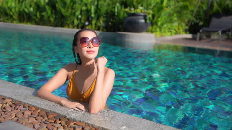 Female-asian-tourist-standing-in-a-swimming-pool-with-half-of-her-body-submerged-in-the-waterduring-a-sunny-day,-wearing-big-sunglasses-and-yellow-swimsuit