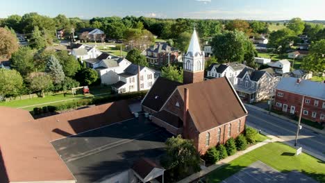Christian-religion-in-America,-United-States-small-town-with-old-brick-church-and-steeple-along-street-with-neighborhood-houses-during-summer,-aerial-orbit-shot