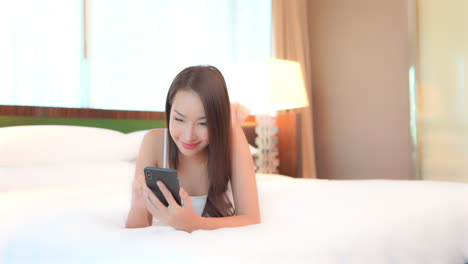 Attractive-girl-lying-on-a-white-sheet-of-her-bed-in-a-hotel-bedroom,-using-her-mobile-phone-and-typing