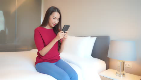 Asiatische-Frau,-Die-Ihr-Telefon-Mit-Beiden-Händen-Benutzt-Und-Auf-Dem-Geschminkten-Bett-Eines-Modernen-Hotelzimmers-Sitzt-Und-Lässige-Kleidung-Trägt