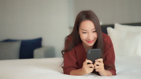 Pretty-Asian-woman-using-her-phone-with-both-hands-typing-laying-on-the-clean-bed-at-the-Hotel-room-wearing-casual-clothes-Close-up-Slow-motion