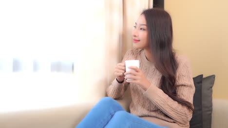 Young-Satisfied-Asian-Woman-Drinking-Coffee-or-Team-by-Window-ay-Sofa-in-Living-Room-of-Family-Home,-Static-Full-Frame-Slow-Motion