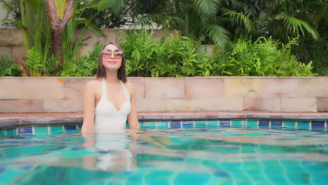 A-pretty-young-woman-in-a-white-one-piece-swimming-suit-enjoys-sitting-in-the-shallow-water-of-a-resort-pool
