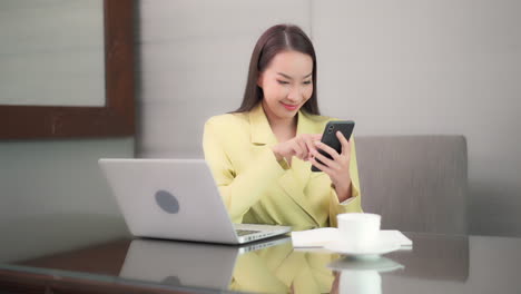 Asiatische-Geschäftsfrau,-Die-Am-Schreibtisch-Vor-Ihrem-Computer-Sitzt-Und-Auf-Dem-Bildschirm-Des-Mobiltelefons-Tippt