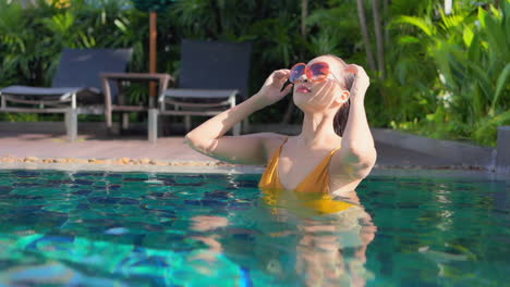 Asian-girl-standing-in-a-swimming-pool,-wearing-a-yellow-bikini-and-sunglasses-correcting-her-wet-hair-and-looking-into-the-camera-afterwards