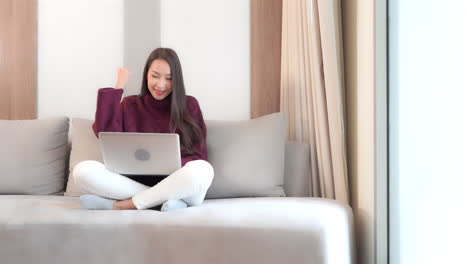 Feliz-Mujer-Asiática-Trabajando-En-Una-Laptop-En-Una-Sala-De-Estar-Con-Un-Gesto-De-Mano-Ganador