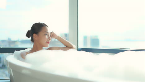 An-Asian-woman-comfortably-taking-bath,-she-is-lying-in-a-bathtub-full-of-foam-at-a-high-floor-of-the-luxury-apartment-complex-with-Glass-walls-in-the-background