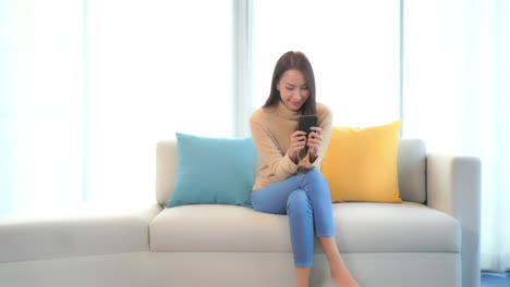 Junge-Asiatische-Frau,-Die-Auf-Der-Couch-Sitzt-Und-Mit-Einem-Lächeln-Auf-Den-Smartphone-Bildschirm-Blickt