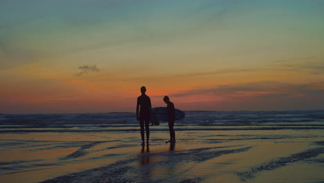 Two-surfer-silhouettes-on-vibrant-beach-during-rainbow-sunset