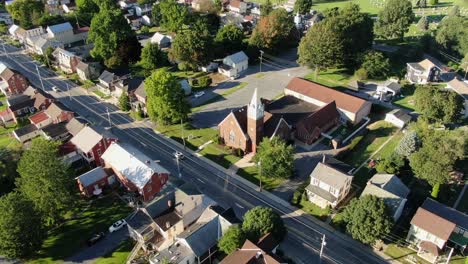 Aerial-orbit-of-small-town,-village-in-United-States,-homes-and-church-during-summer-magic-hour-as-traffic-passes-by,-drone-shot