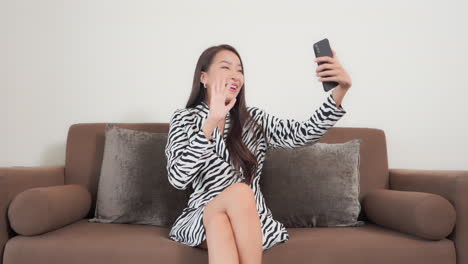Asian-woman-having-a-lovely-video-call-conversation-using-her-mobile-phone-camera-sitting-on-the-sofa-at-home,-girl-waves-palm-and-says-hello-to-the-camera,-shows-OK-gesture