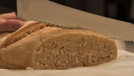 Female-hand-holding-and-cutting-through-freshly-baked-loaf-of-sour-dough-bread-with-sharp-serated-kitchen-knife-on-benchtop