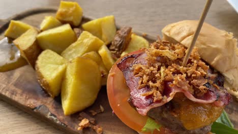 Mini-burger-with-bacon,-crispy-onions,-tomato,-lettuce-and-potato-cubes-on-the-side-in-a-restaurant-on-a-wooden-platter,-4K-panning-left