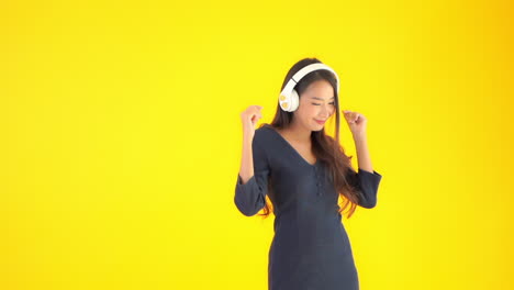 Una-Encantadora-Chica-Asiática-Que-Usa-Auriculares-Bluetooth-En-La-Oreja-Está-Escuchando-Música-Y-Bailando-En-Un-Estudio-Amarillo