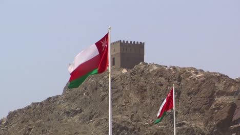 Zwei-Im-Wind-Wehende-Flaggen-Des-Staates-Oman-Im-Hintergrund-Sieht-Man-Den-Turm-Der-Festung