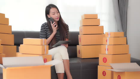 A-young-pretty-businesswoman-surrounded-by-packages-ready-for-shipping-checks-logistics-on-her-iPad-while-answering-her-smartphone