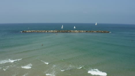 Olas-Rodando-En-La-Playa-Con-Veleros-Navegando-Más-Allá-Del-Malecón-Junto-Al-Mar-Mediterráneo-En-Tel-Aviv,-Israel