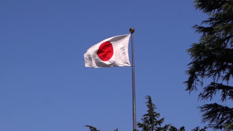 Mittlere-Aufnahme-Der-Japanischen-Flagge-Auf-Flaggenmast,-Die-In-Zeitlupe-Gegen-Den-Blauen-Himmel-Mit-Baumsilhouetten-Weht